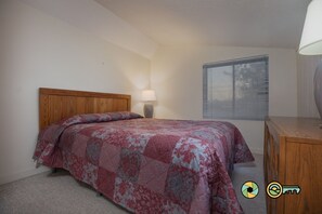 A bedroom with a wooden headboard, a patterned bedspread, a bedside lamp, and a window with blinds.