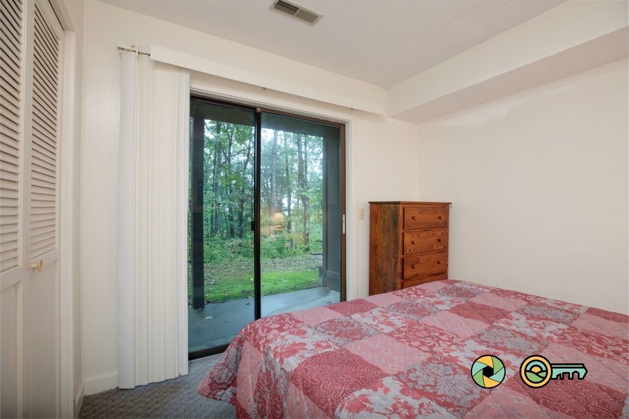 A bedroom with a bed, a wooden dresser, and a sliding glass door leading to a balcony with a view of trees.