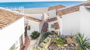 Un bâtiment situé sur la côte, doté d’un toit recouvert de tuiles en terre cuite, de murs blancs et d’un petit jardin agrémenté de diverses plantes et d’un élément décoratif en roche.