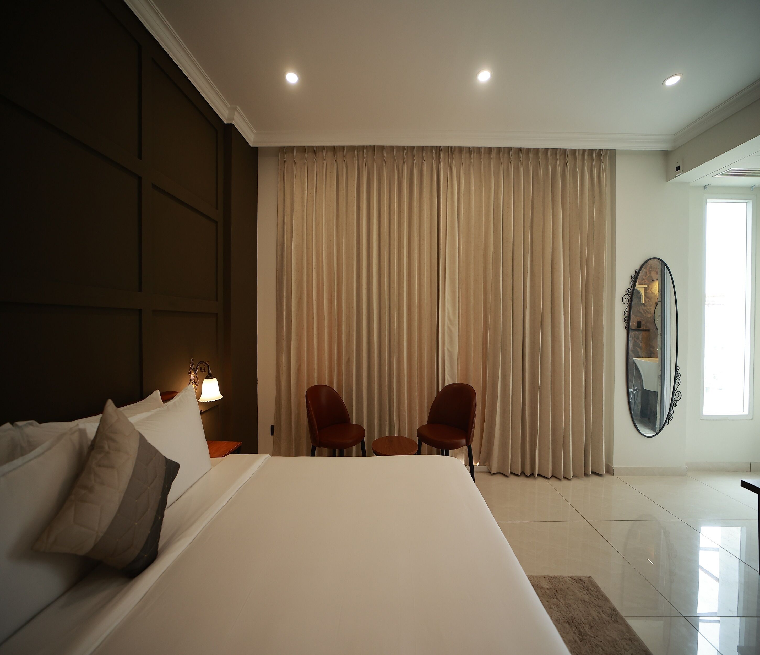 A hotel room with a large bed, two chairs, a mirror, and a window with curtains.