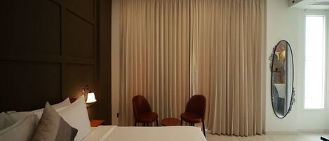A hotel room with a large bed, two chairs, a mirror, and a window with curtains.