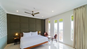 A hotel room with a large bed, a ceiling fan, a nightstand with a lamp, a chair, a small table, and a balcony with curtains. - De Fleur Hotel (Puducherry)