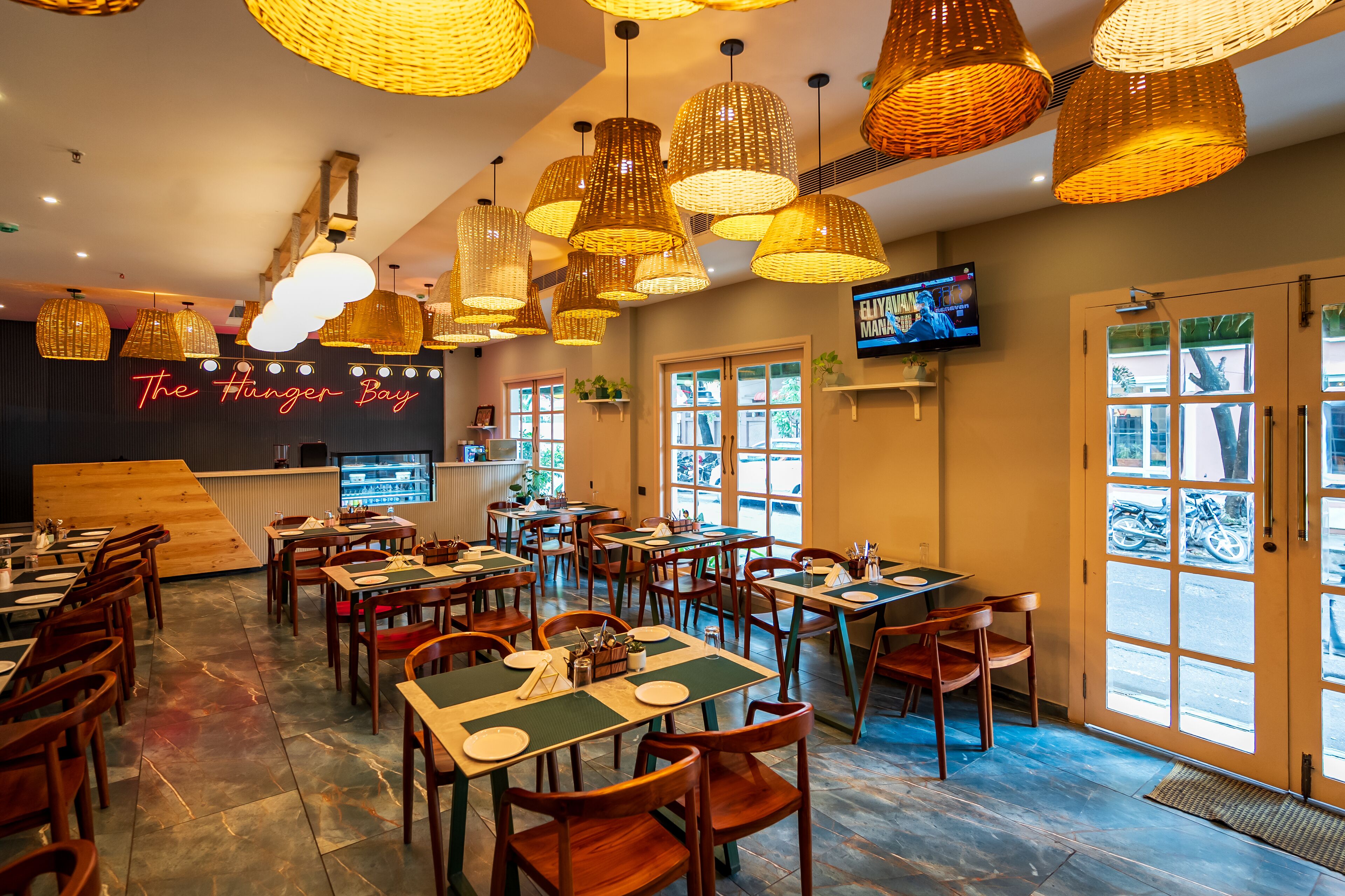A restaurant interior with multiple tables and chairs, hanging wicker lamps, and a television on the wall.