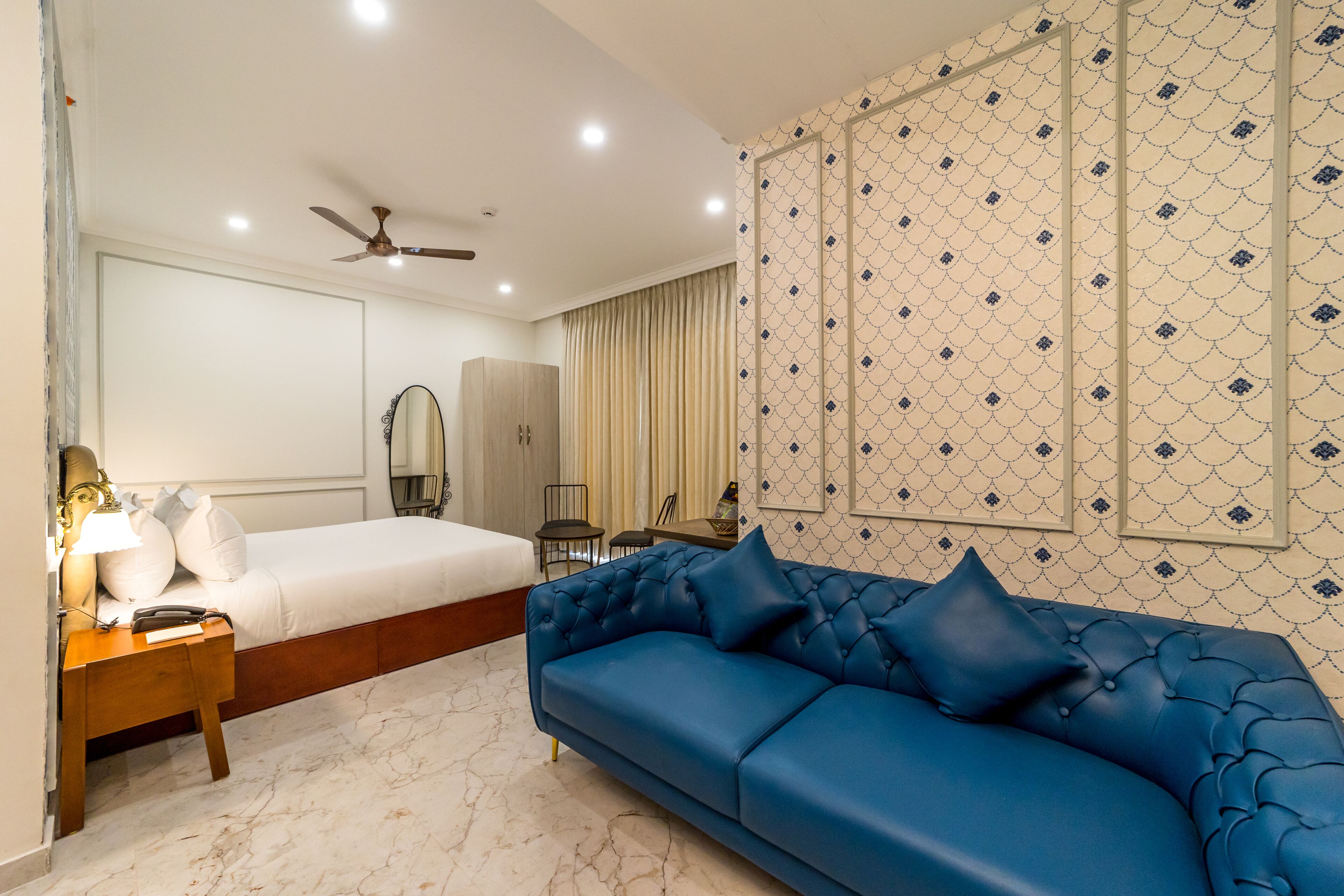A hotel room with a bed, a blue sofa, a nightstand, a mirror, a wardrobe, and a ceiling fan.