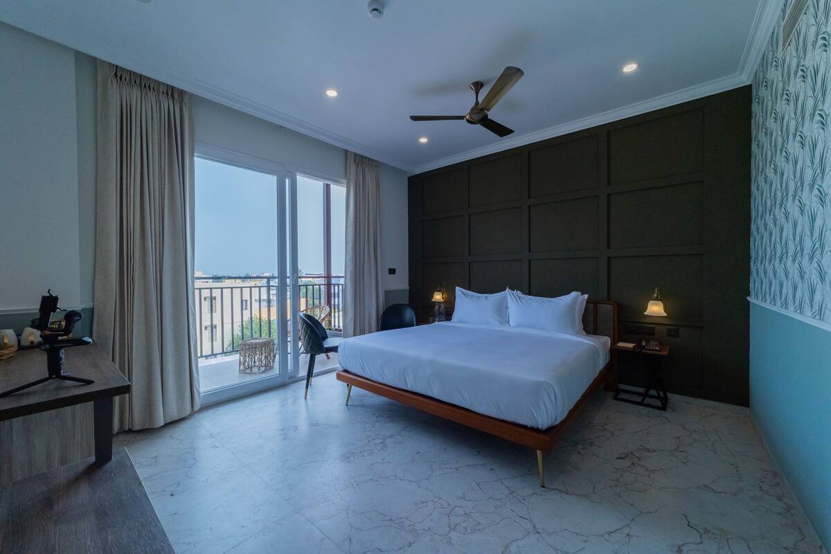 A hotel room with a large bed, a desk, a chair, a balcony with a view, and a ceiling fan.