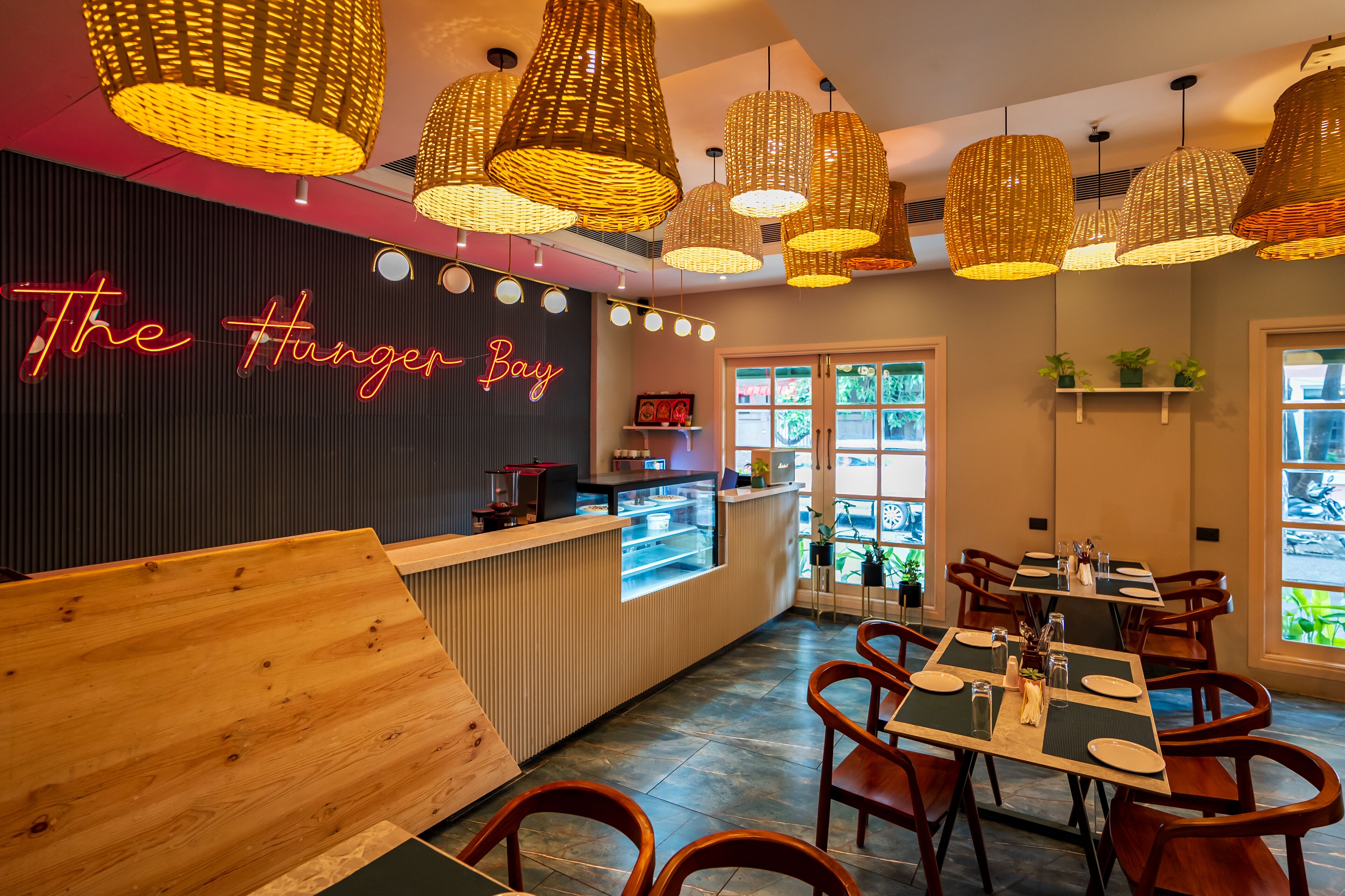 A restaurant interior with a neon sign reading "The Hunger Bay," wooden reception desk, wicker pendant lights, and a dining area with tables and chairs.