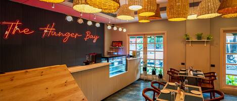 A restaurant interior with a neon sign reading "The Hunger Bay," wooden reception desk, wicker pendant lights, and a dining area with tables and chairs.