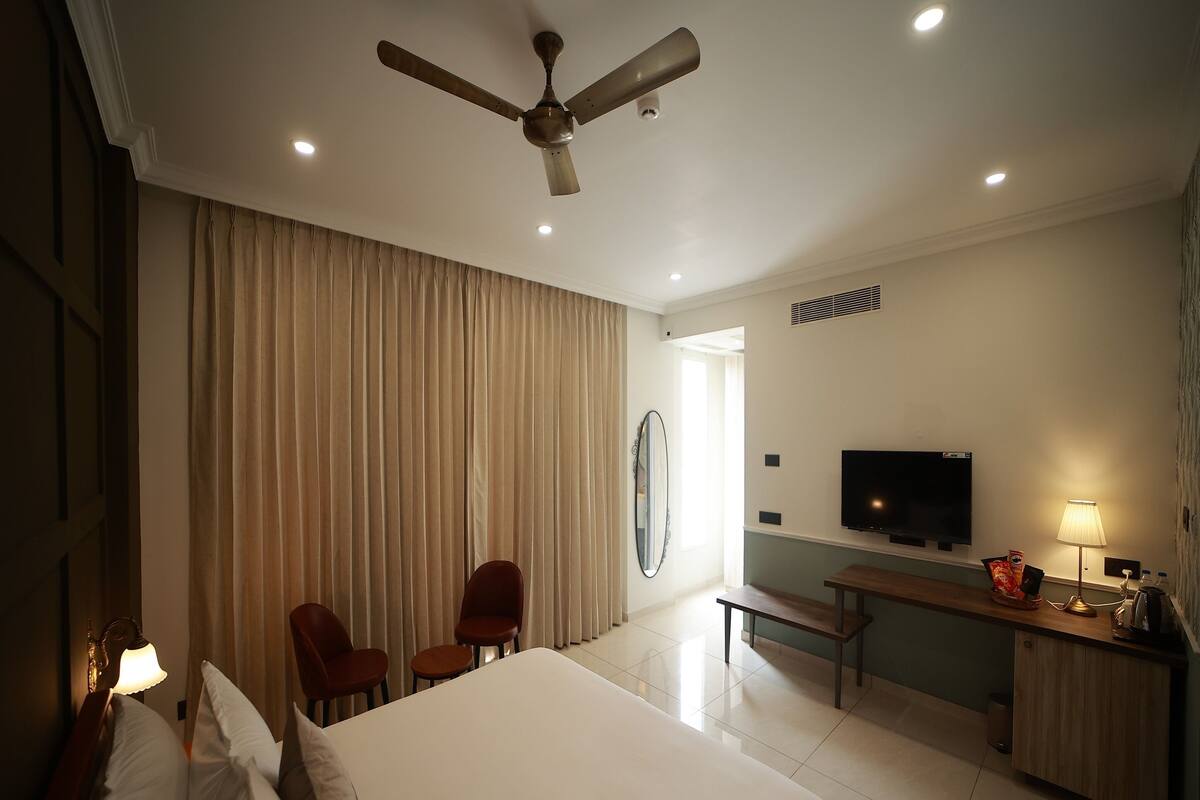 A hotel room with a bed, two chairs, a desk with a TV, a lamp, and a ceiling fan.