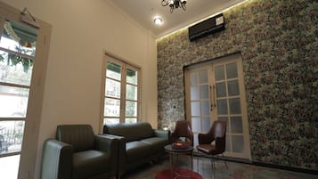 A living room with a sectional sofa, two armchairs, a small table with a bowl of fruit, a red rug, and a patterned wallpaper.