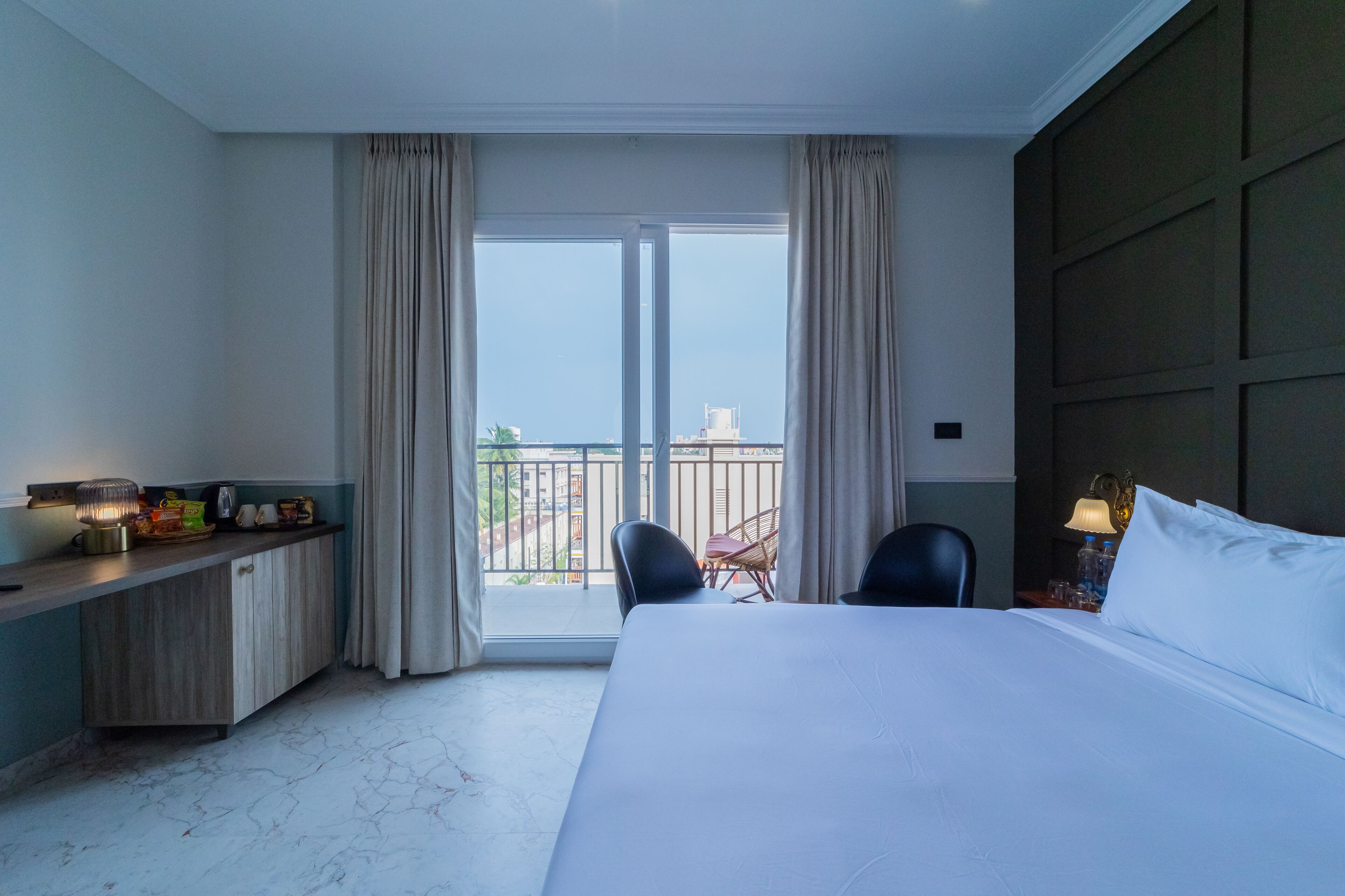 Habitación de hotel con cama, escritorio con lámpara, una silla y vistas al exterior.