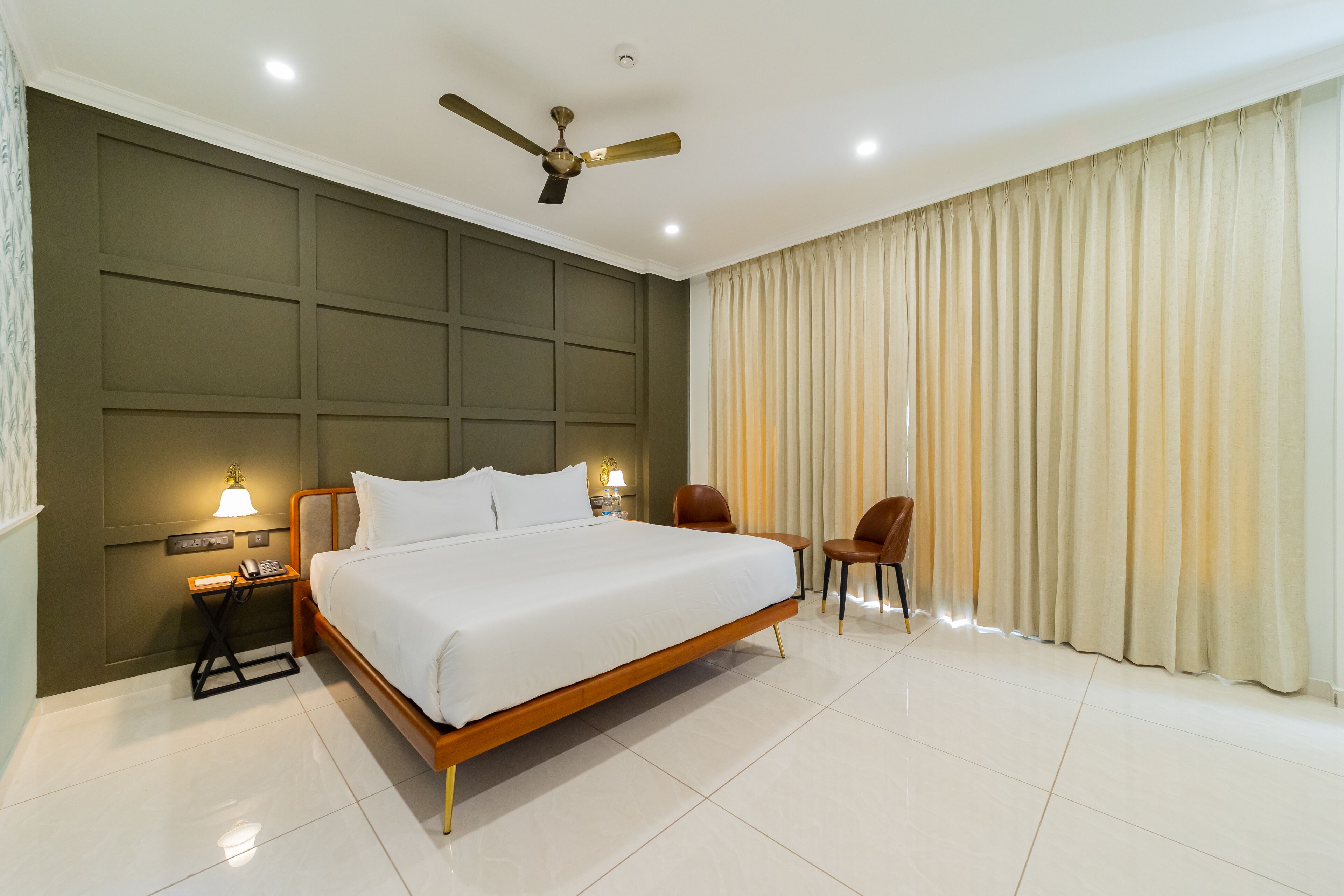 A hotel room with a large bed, a desk with a lamp, two chairs, a ceiling fan, and a large window with curtains.