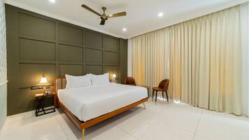 A hotel room with a large bed, a desk with a lamp, two chairs, a ceiling fan, and a large window with curtains.