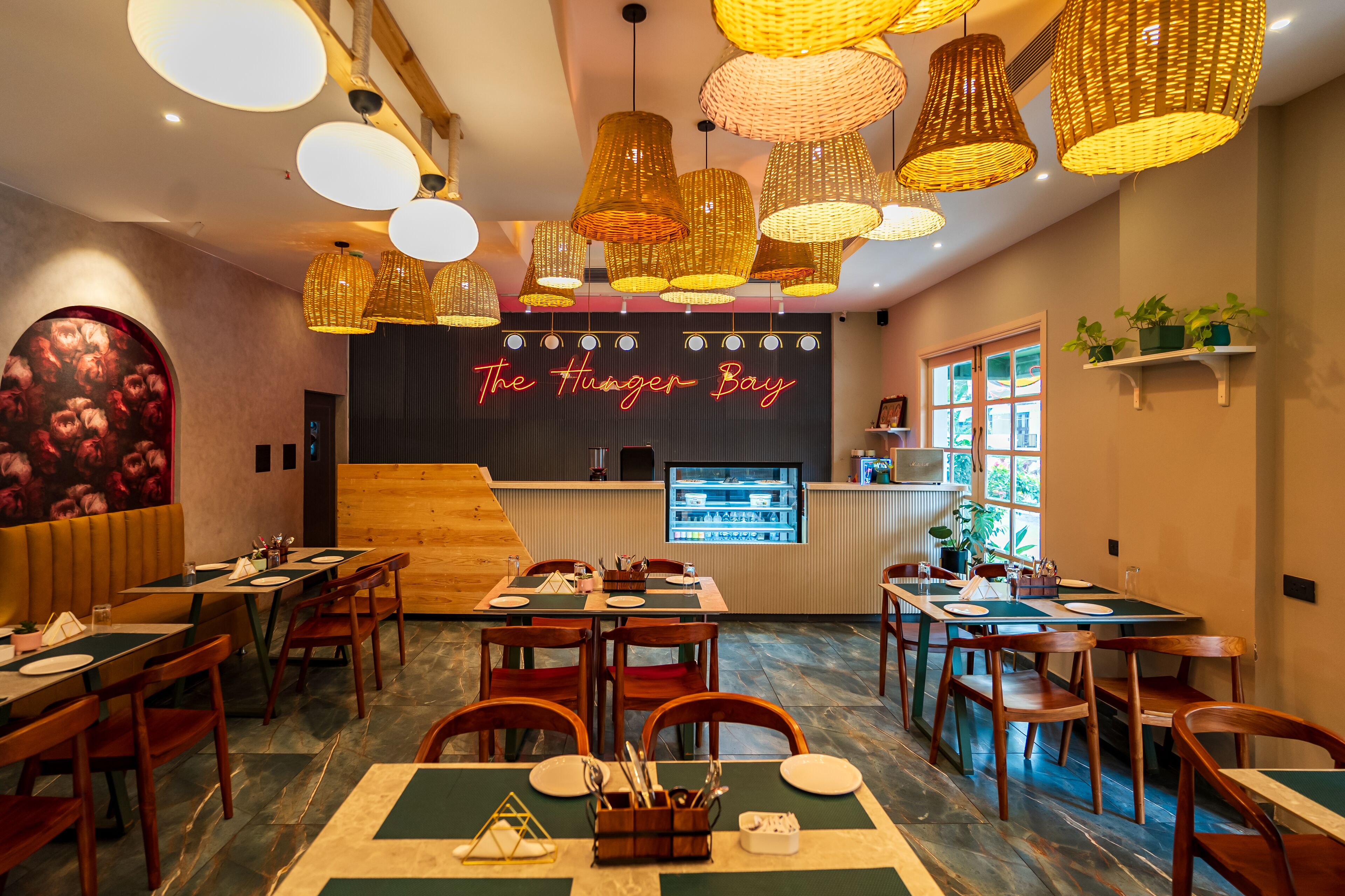 A restaurant interior with wooden tables and chairs, hanging wicker lamps, and a neon sign reading "The Hunger Bay".