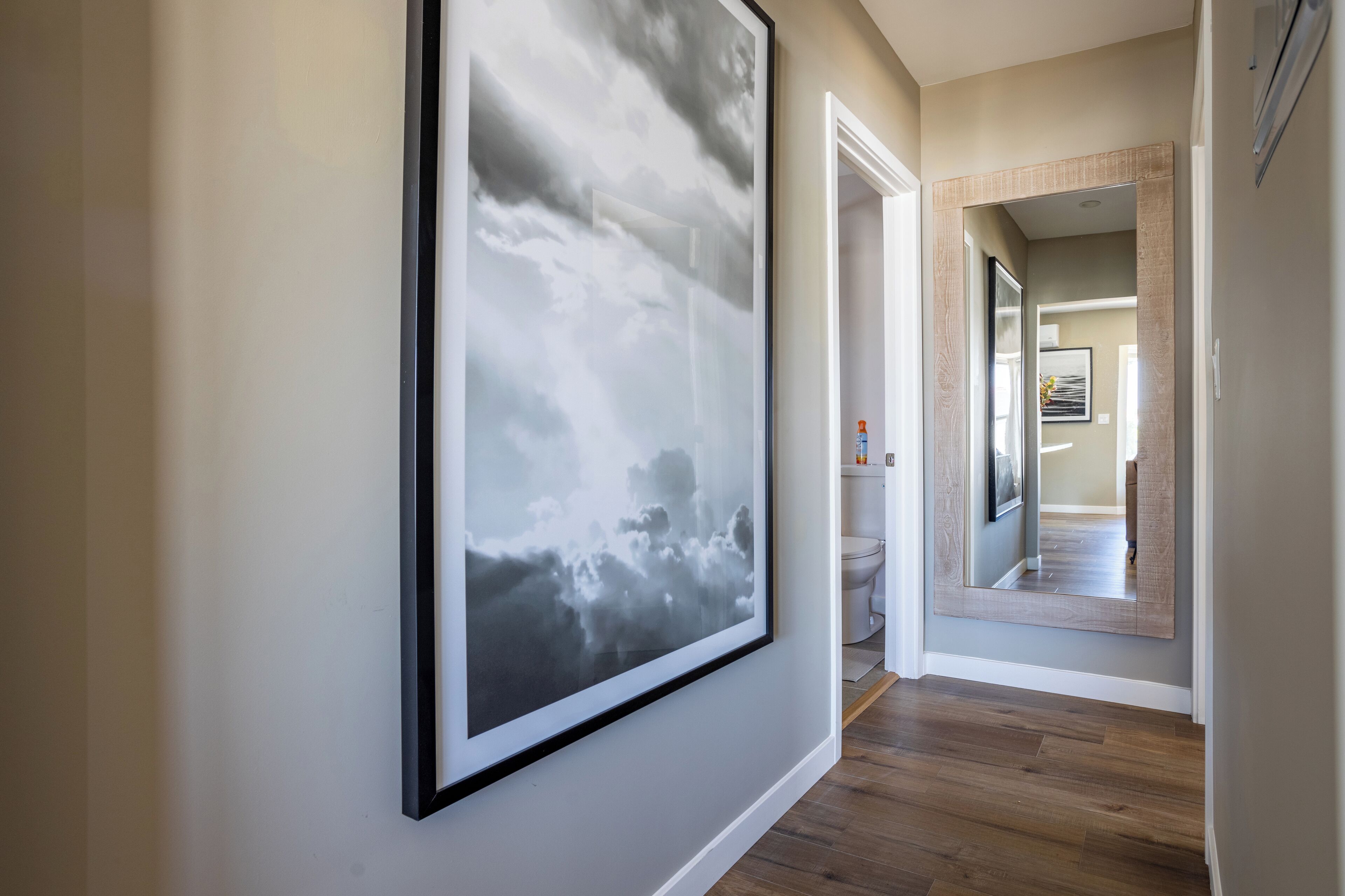 A hallway with a framed abstract painting, a wooden floor, and a mirror reflecting a bathroom with a toilet and a sink.