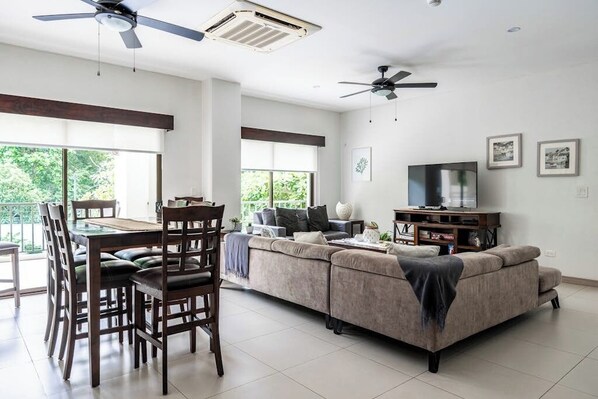 Ein geräumiges Wohnzimmer mit Sofa, Esstisch, Stühlen, Deckenventilatoren und einem Fernseher.