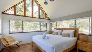 Een slaapkamer met een groot bed, een houten hoofdbord en uitzicht op bomen door de ramen.
