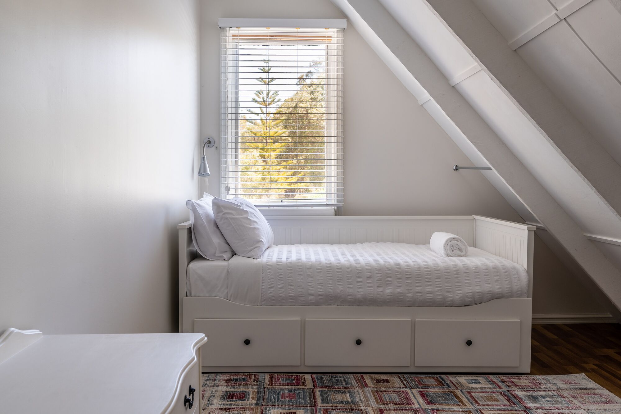 Een kleine, witte slaapkamer in loftstijl met een schuine kap, een raam met jaloezieën, een bed met opbergladen en een kamerbreed tapijt met dessin.