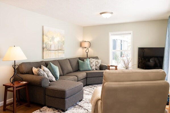 A living room with a sectional sofa, a chair, a lamp, a painting, and a television.