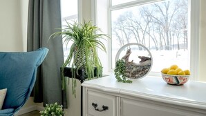 A room with a blue sofa, a white cabinet, a potted plant, a decorative bowl with lemons, and a view of trees through a window.
