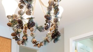 A dining table with a clear glass vase containing a plant, white chairs, and a chandelier with pine cones.