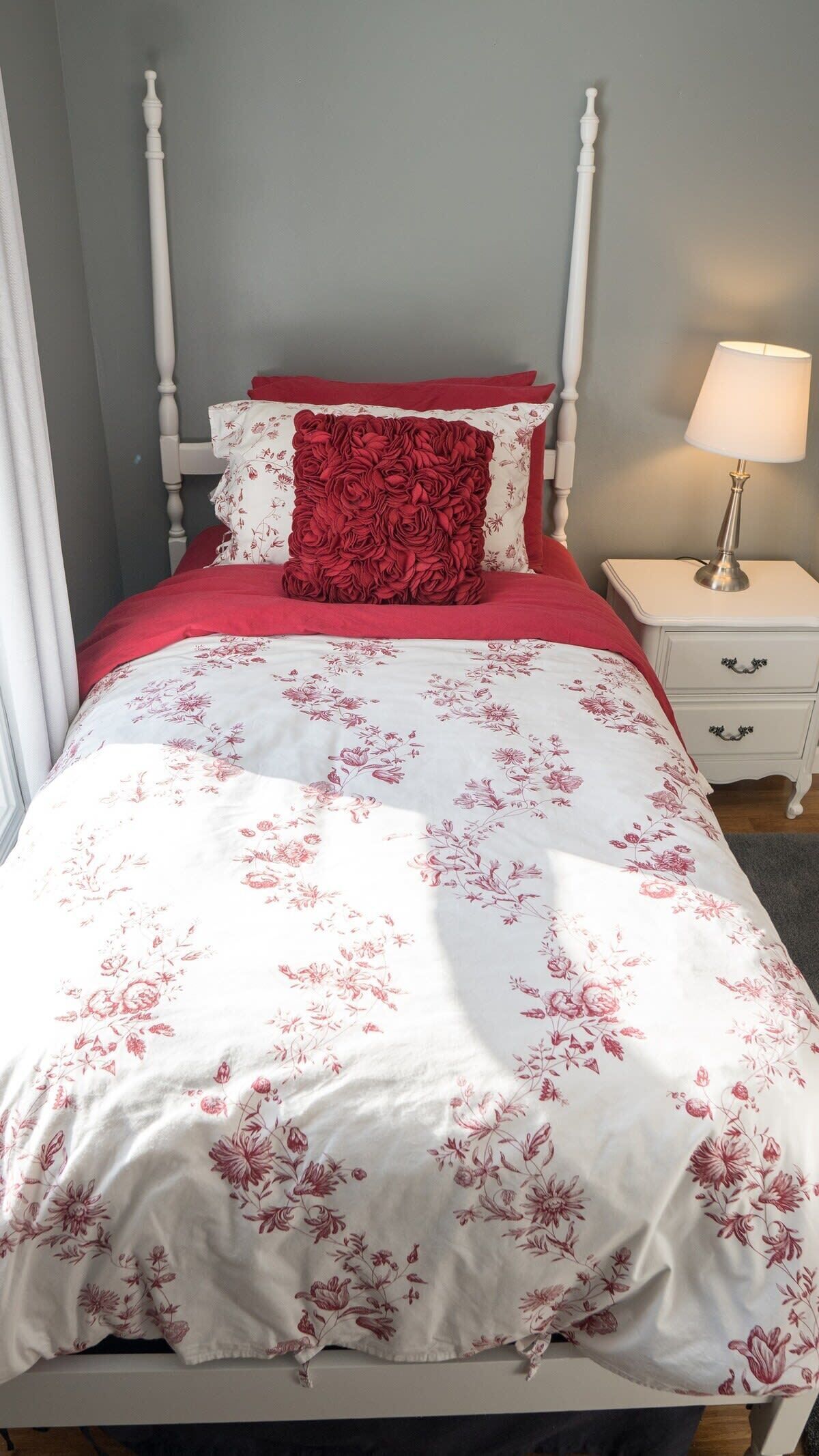 A bed with a white and red floral bedspread, a red pillow, and a white nightstand with a lamp.
