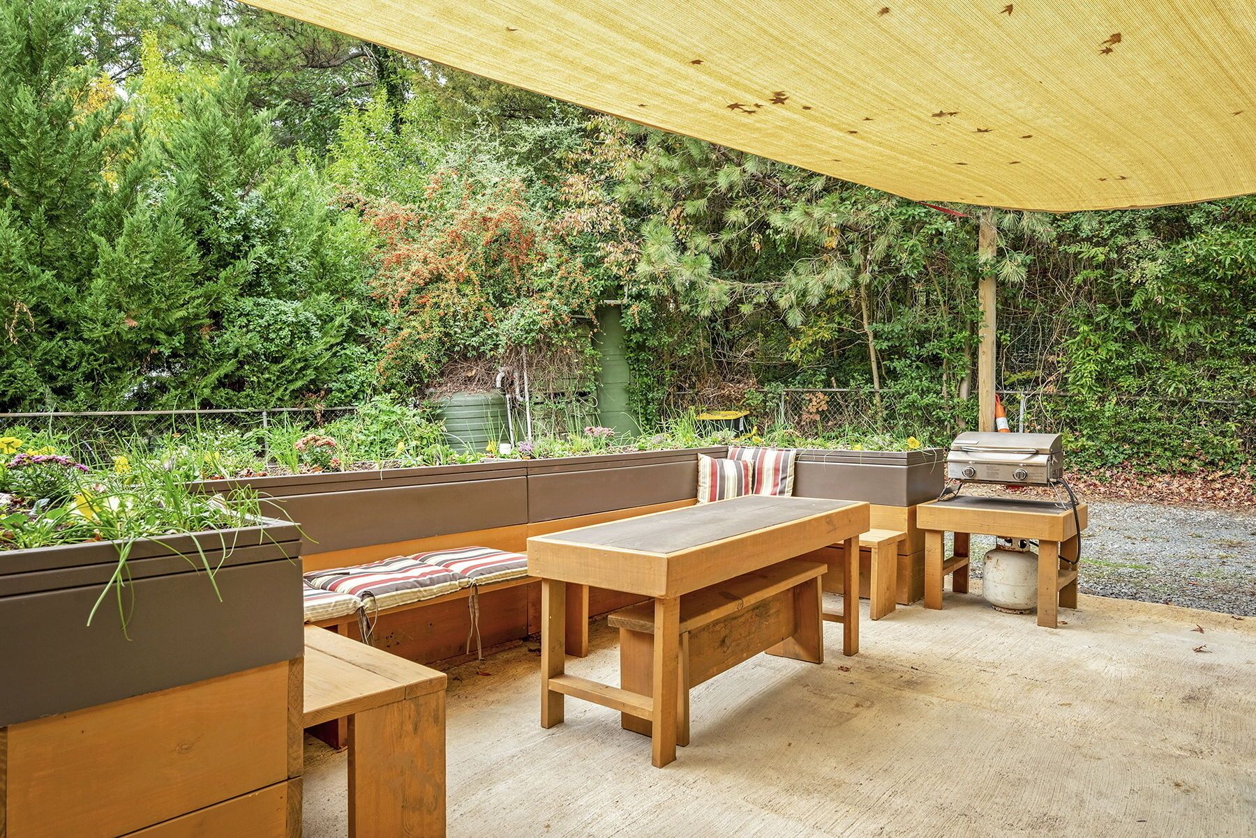 Un área de estar al aire libre con bancos y mesas de madera, una parrilla para barbacoa y un toldo grande.