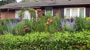 Une maison avec une façade en briques, de grandes fenêtres et un jardin où poussent différentes plantes et fleurs.