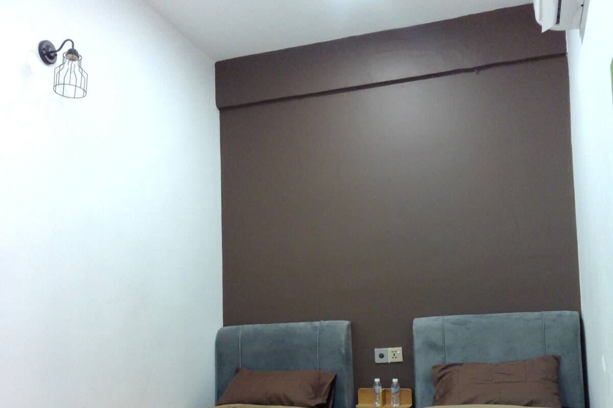 Una stanza piccola e modesta con due letti singoli, un comodino con bottiglie d'acqua e un lampadario a parete.