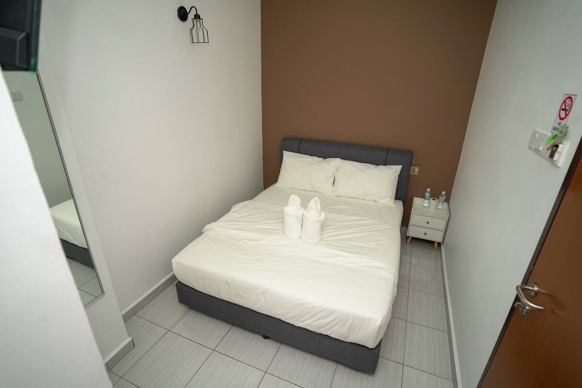 Letto ben rifatto con lenzuola bianche e due asciugamani, un comodino con una lampada e un interruttore a parete.