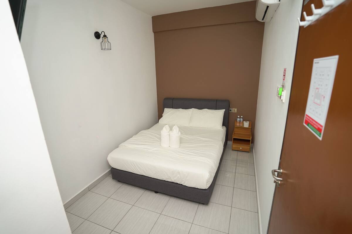 Un letto rifatto con lenzuola bianche e due asciugamani, un comodino con una lampada, e una porta con un cartello.