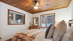 A bedroom with a wooden ceiling, a bed with patterned bedding, a nightstand, a mirror, and windows with a snowy view. - The Lazy Bear - Mountain Retreat with Sauna (Whitefish)