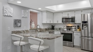 A modern kitchen with white cabinets, stainless steel appliances, a marble countertop, and two bar stools. - Tampa's Paradise (Tampa)