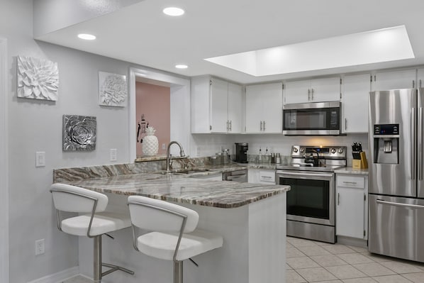 A modern kitchen with white cabinets, stainless steel appliances, a marble countertop, and two bar stools.