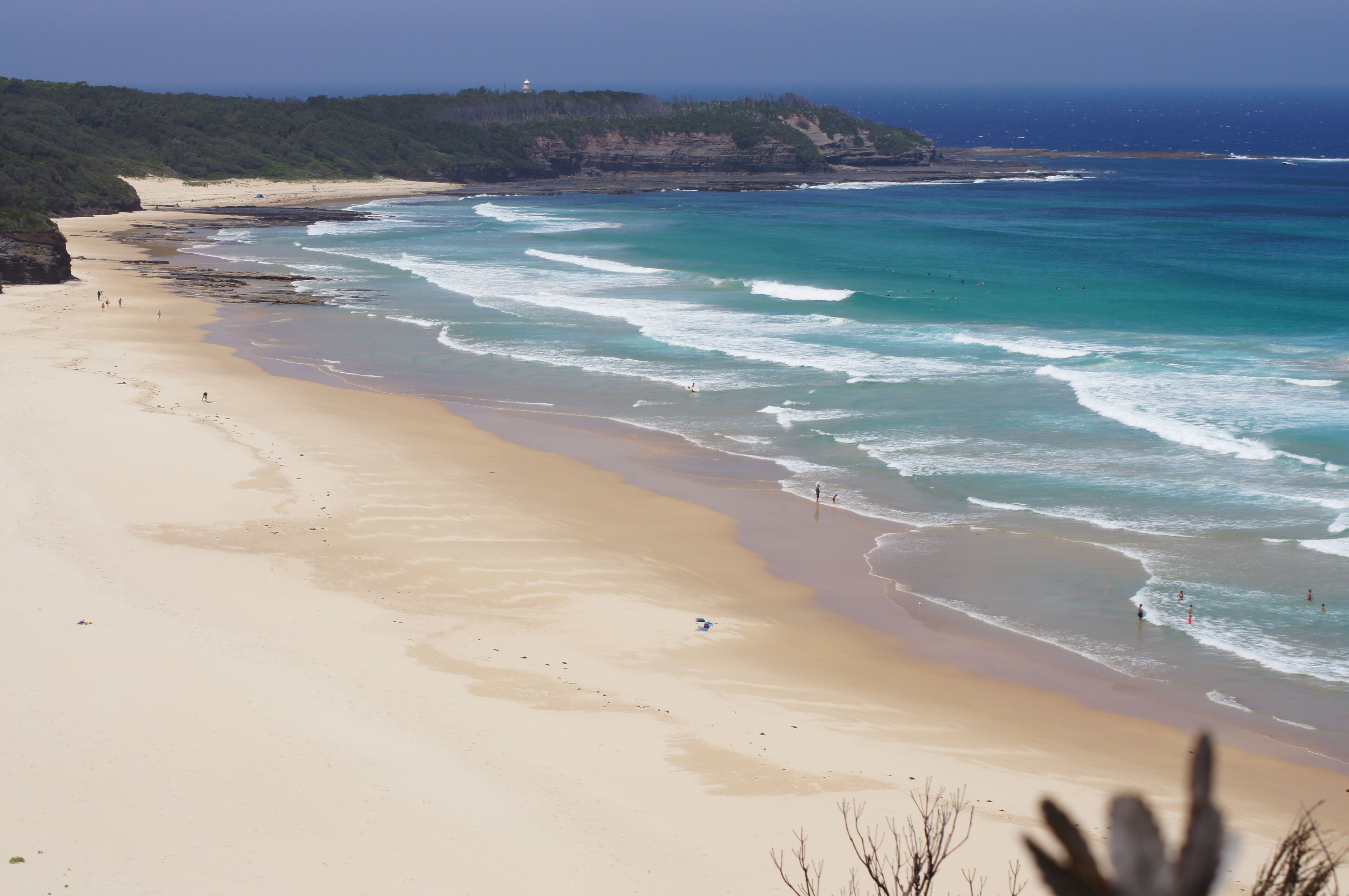Una playa con aguas turquesas cristalinas, una orilla arenosa y un faro a lo lejos.