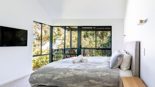A bedroom with a large bed, a TV mounted on the wall, and a balcony with a view of trees. - Surfside Retreat | Gnarabup (Gnarabup)