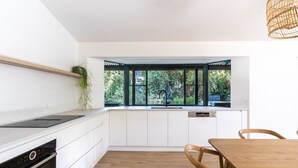 A modern kitchen with white cabinetry, a black induction cooktop, and a window with a view of greenery. - Surfside Retreat | Gnarabup (Gnarabup)