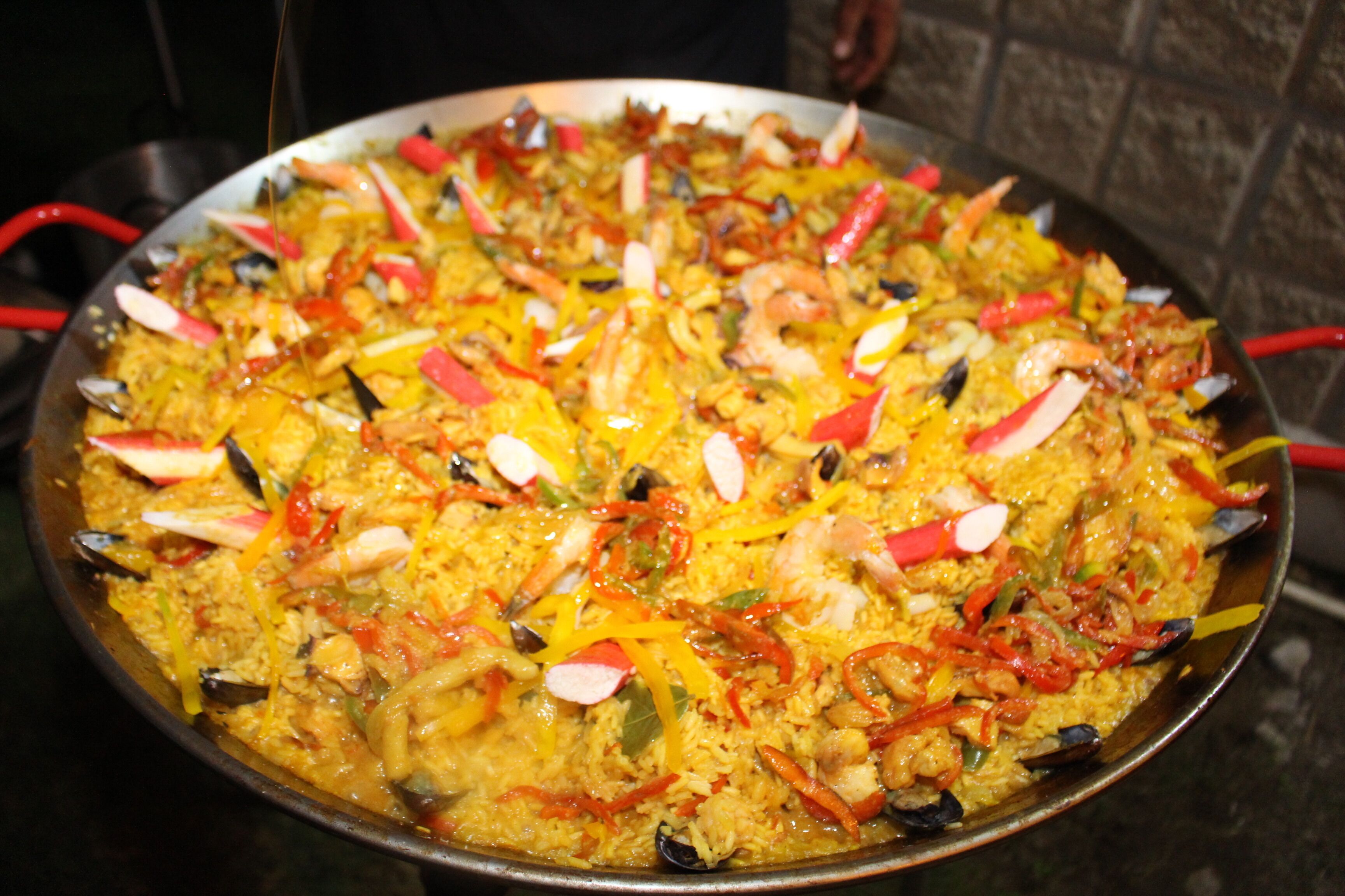 Eine große, runde, flache Pfanne gefüllt mit einem gelben Reisgericht, garniert mit roten Paprika, grünen Zwiebeln und weißen Scheiben, vermutlich mit Meeresfrüchten.