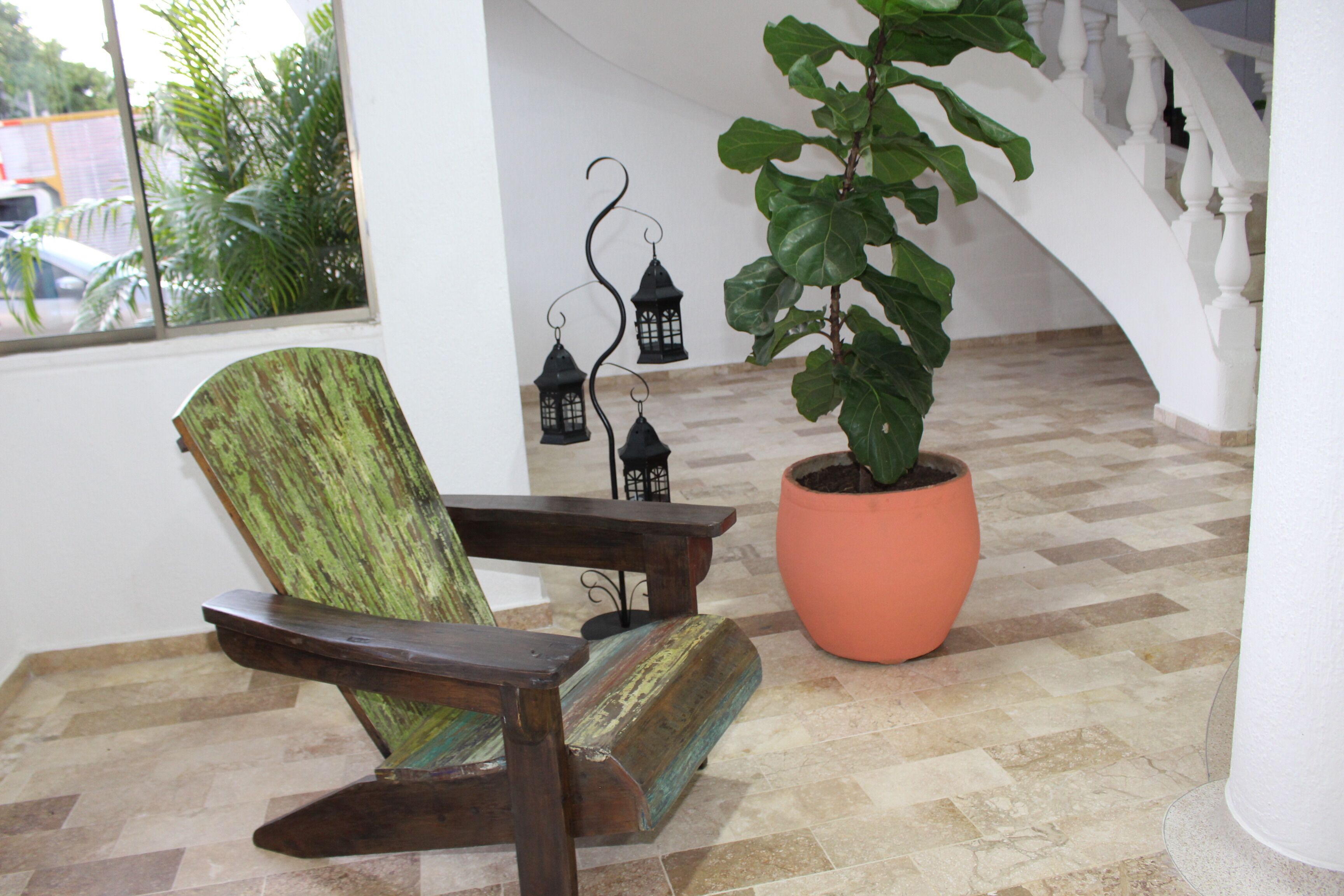 Una silla de madera con acabado envejecido, una planta en maceta y faroles decorativos en una habitación con piso a cuadros y paredes blancas.