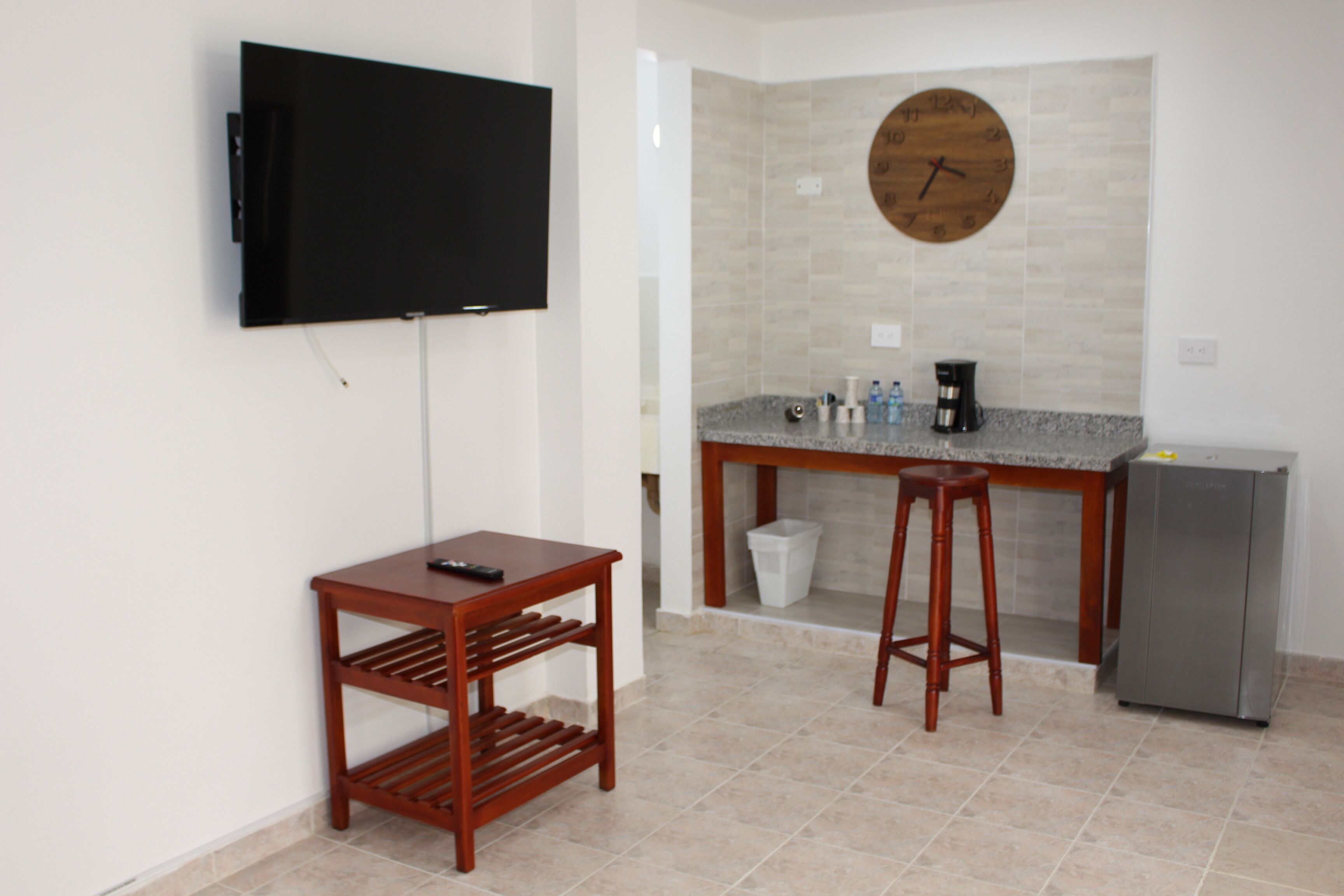 Una cocina pequeña con televisor, una mesa de madera, un taburete de bar, un refrigerador y un reloj en la pared.