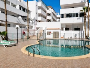 Una piscina rodeada por un edificio blanco con balcones, palmeras y un paseo de azulejos.
