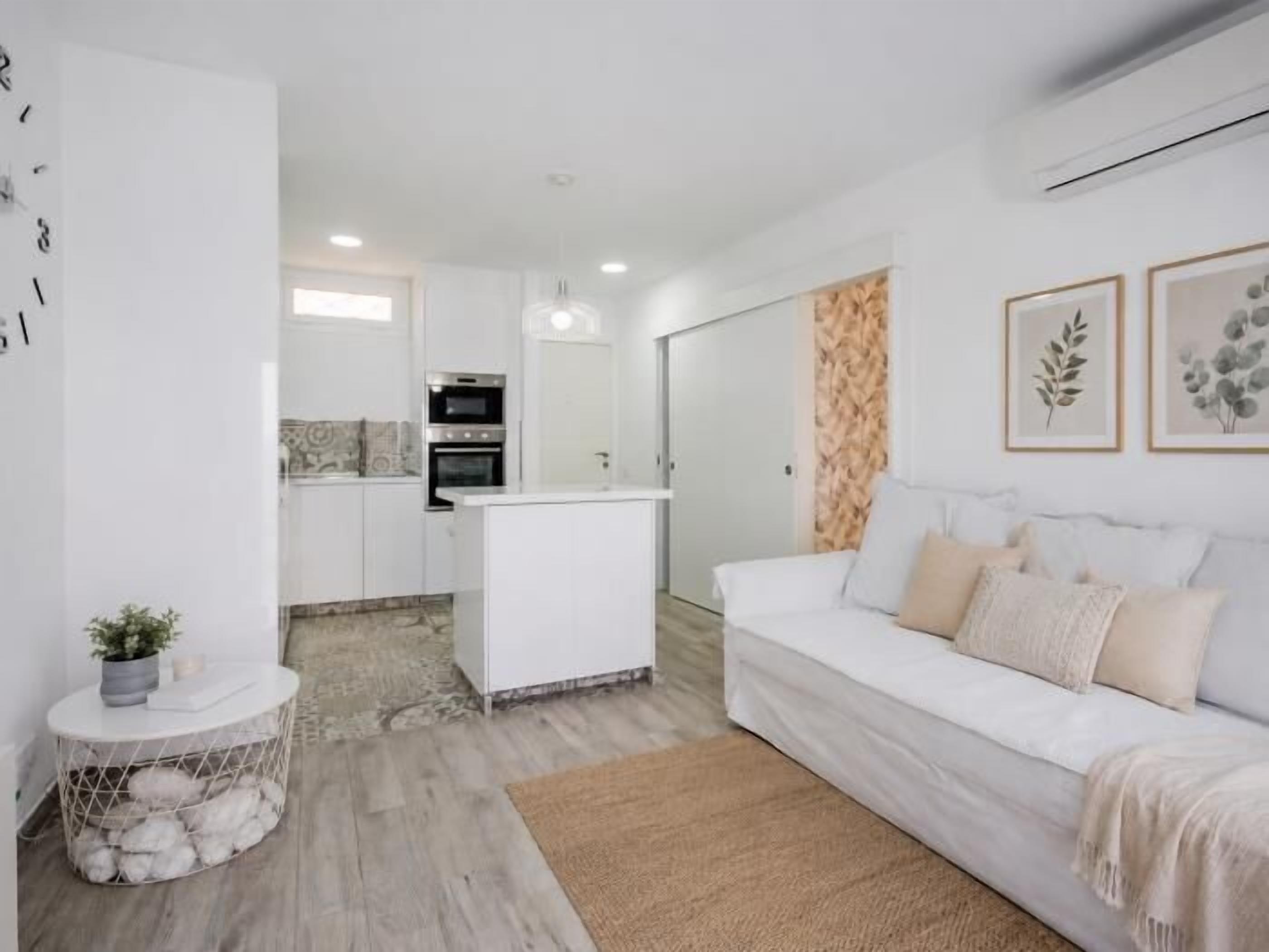 A modern living room with a white sofa, a small round table with a plant, and a kitchen area in the background.