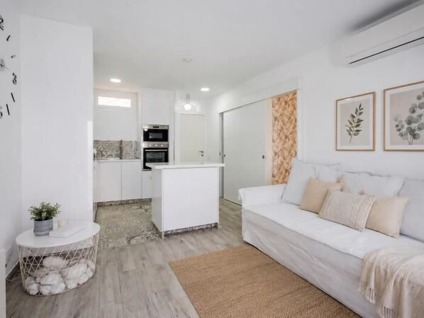 A modern living room with a white sofa, a small round table with a plant, and a kitchen area in the background.
