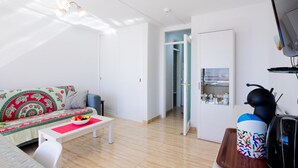 A modern living room with a sofa, a coffee table, and a TV. There is a door leading to another room and a shelf with items on it.