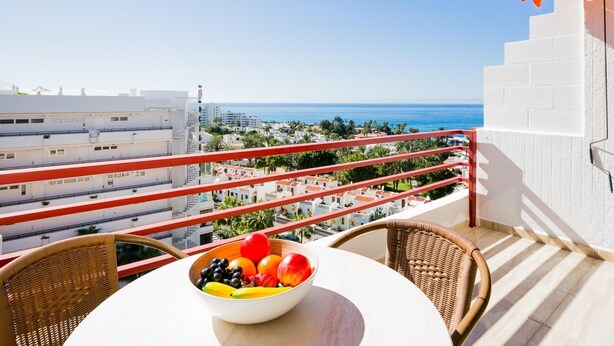 A balcony with a table and chairs, a bowl of fruit, and a view of the sea and buildings.
