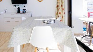 A modern room with a long table, white chairs, a flat-screen TV, a coffee machine, and a window with curtains.
