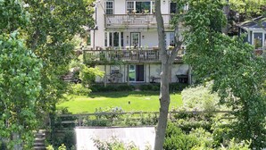 A two-story house with a balcony, surrounded by lush greenery and a well-maintained lawn.