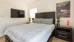 Une chambre à coucher avec un lit, un téléviseur fixé au mur, une table de chevet avec une lampe et un tableau accroché au mur.