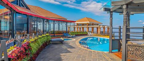 A pool area with a glass-enclosed lounge, a gazebo, and a view of the sea.