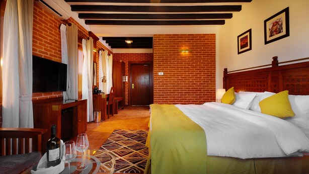 Une chambre d’hôtel avec un mur en briques, un lit en bois avec des oreillers jaunes, une télévision, un bureau avec une chaise et un tapis à motifs.