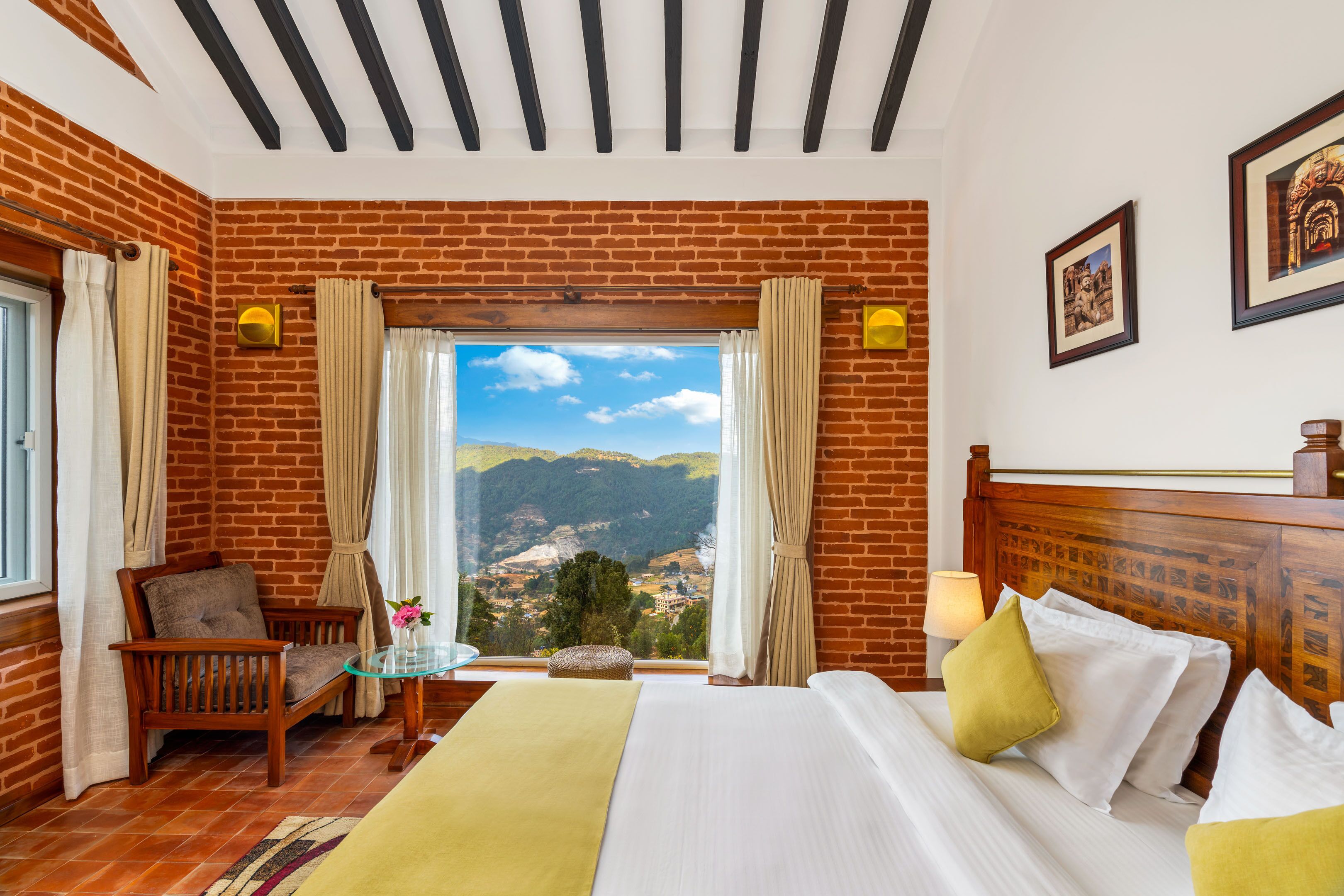 A room with a brick wall, a bed with white bedding, a wooden headboard, a chair, a small table with a glass top, and a view of the countryside through a window.